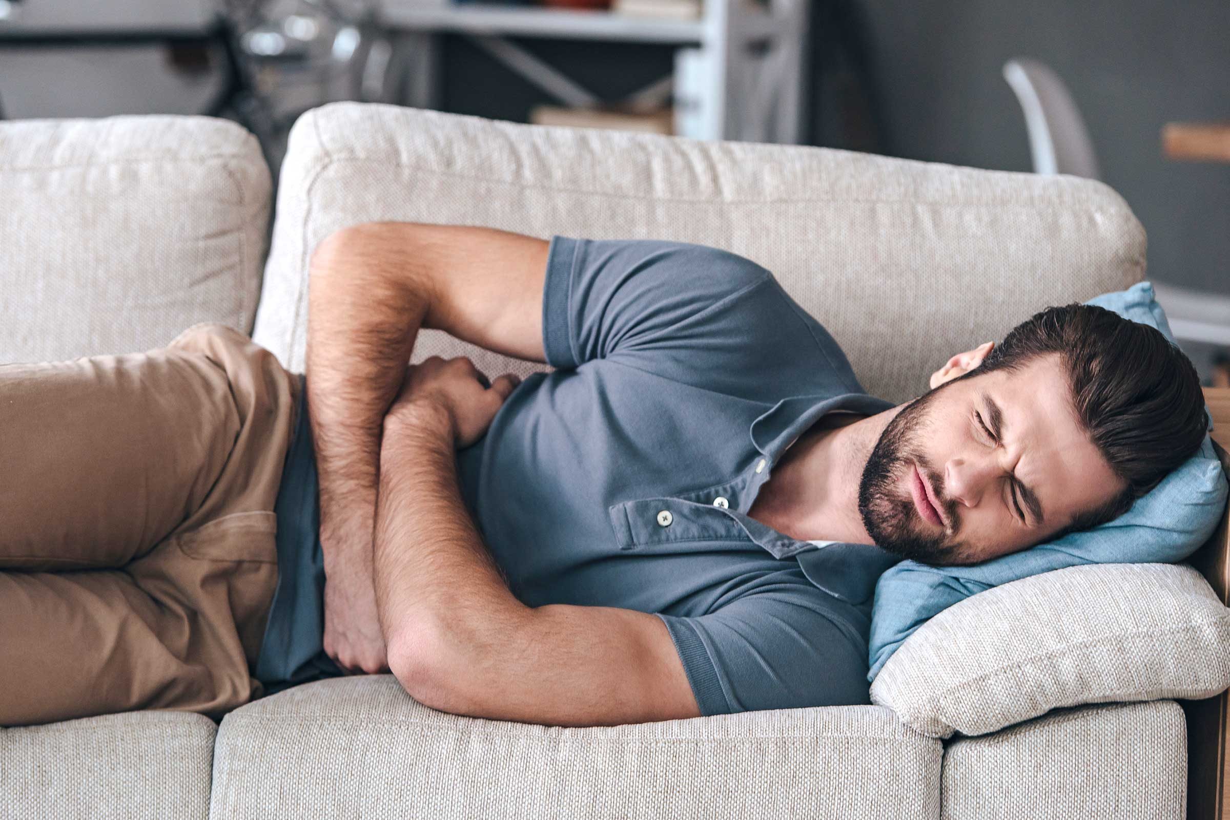 bearded man on his side on a couch, grimacing and holding his stomach