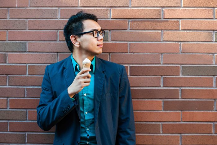 Man in a suit eating an ice cream cone in front of a brick wall.