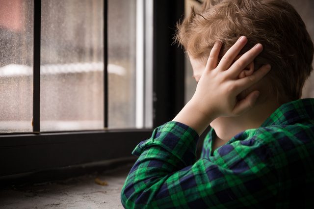 young boy sitting near window with hands over ears