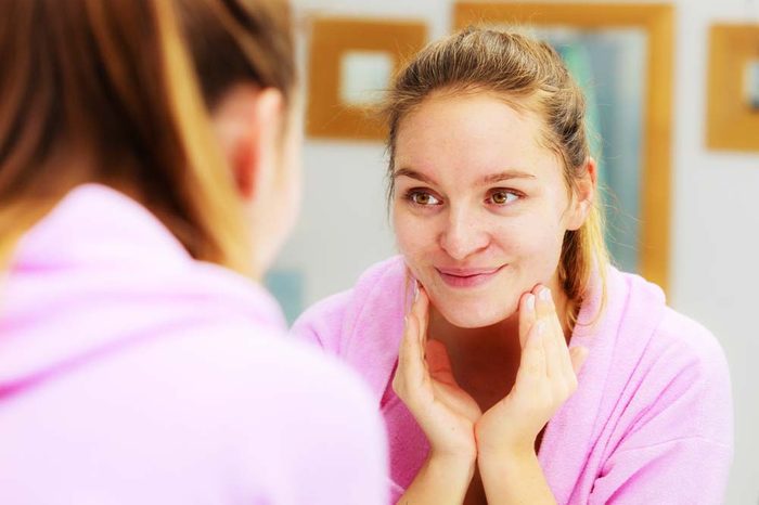 Woman wearing a pink hoodie looking at her skin in the mirror.