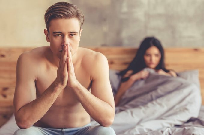 Young couple having a problem. Man is holding hands together while sitting on bed, woman is lying in the background