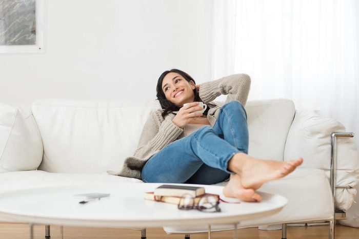 Young smiling woman sitting on sofa and looking up while drinking hot tea. Young brunette woman thinking at home in a leisure time. Happy girl relaxing at home on a bright winter morning.