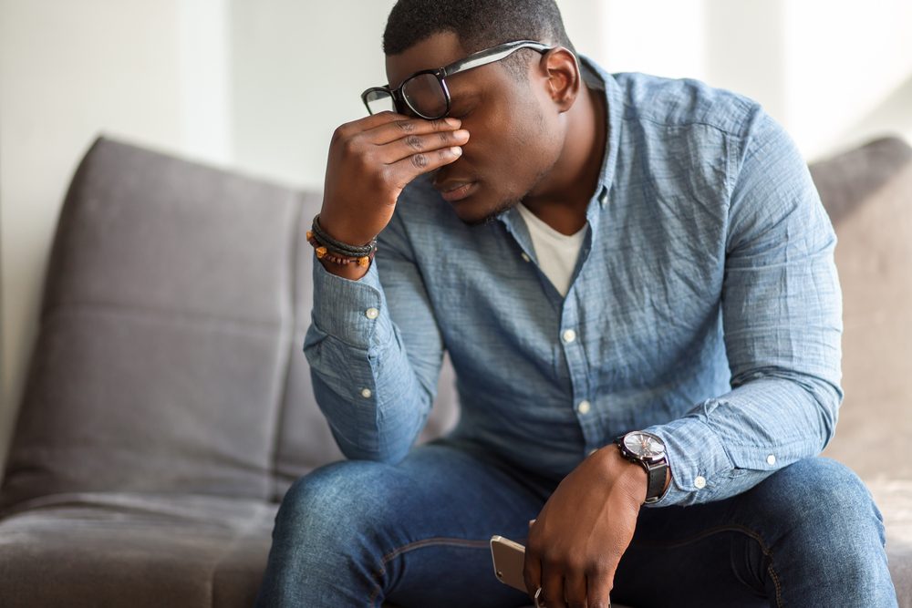 Feeling exhausted. Frustrated young black man massaging his nose and keeping eyes closed while sitting at his working place in office