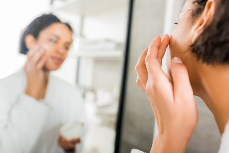 selective focus of african american woman applying cosmetic cream on face in bathroom