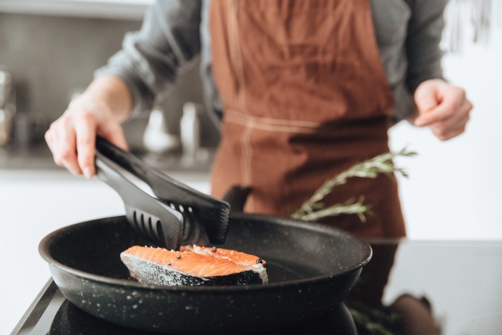 Cropped image of young lady standing in kitchen while cooking salmon.