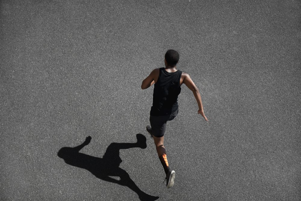 Top view of running man sprinting for success on run. Muscular runner or jogger dressed in black outfit, training at fast speed on black asphalt. Fit sport model exercising sprint outdoors