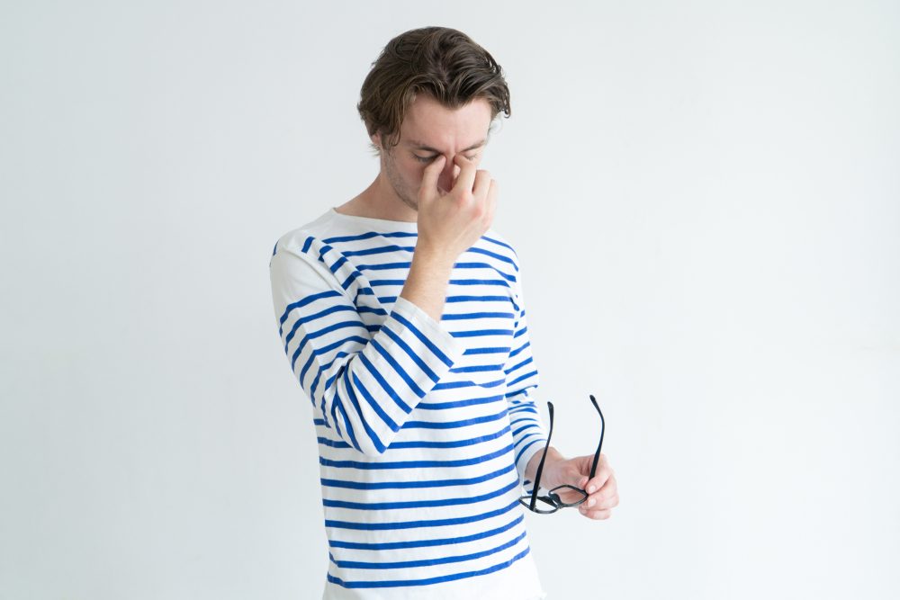 Exhausted man rubbing bridge of nose while feeling eye fatigue. Tired young male manager massaging nose and holding glasses. Tension concept