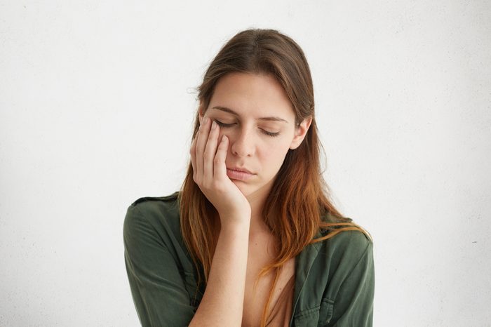 Woman with sleepy expression holding her hand on cheek, eyes closed. 