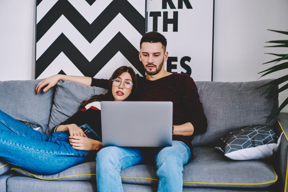 Pensive young marriage couple resting on cozy couch and watching movie on laptop computer. Lovely female and male spending time lounge in apartment and using internet at home on lounge sofa