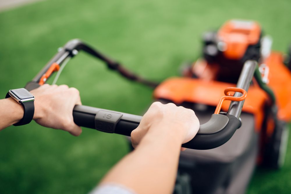A man holds a lawn mower for the handle. He came to the tools store for gardening. He has a wristwatch on his hand.