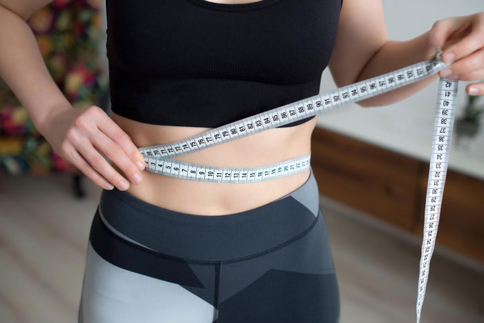 A young woman in a sportswear checking success of weight loss program with measuring tape in front of mirror at home.