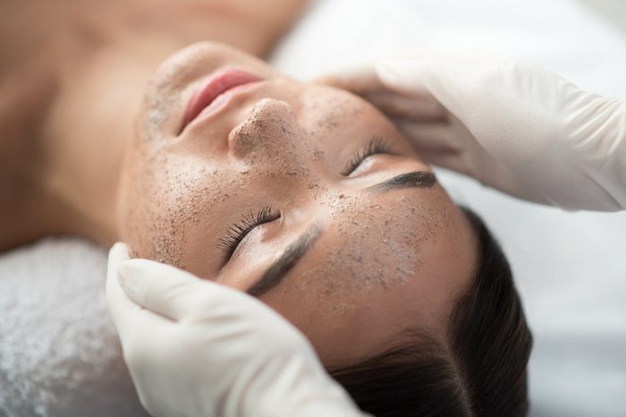 Close up of young woman getting facial skin treatment at spa. Cosmetician hands applying peeling mask on her face