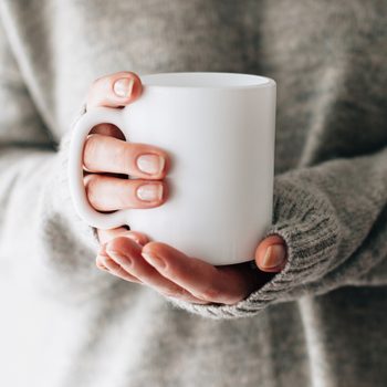 Closeup of female hands with a mug of beverage. Beautiful girl in grey sweater holding cup of tea or coffee in the morning sunlight. Mug for your design. Empty.