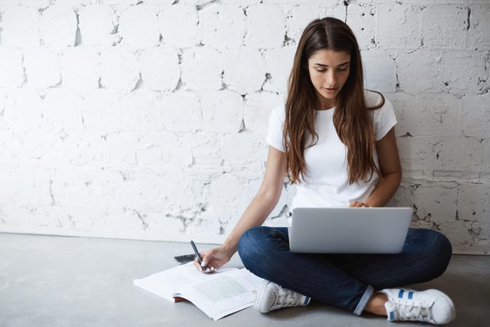 Indoor portrait of smart busy woman, making notes while leaning on wall and sitting on floor with laptop on crossed feet. Girl has habit to work or study everywhere but not at table. Study and work
