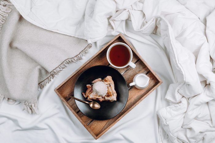 Cozy flatlay of bed with wooden tray with vegan apple pie, ice cream and black tea on white sheets and blankets