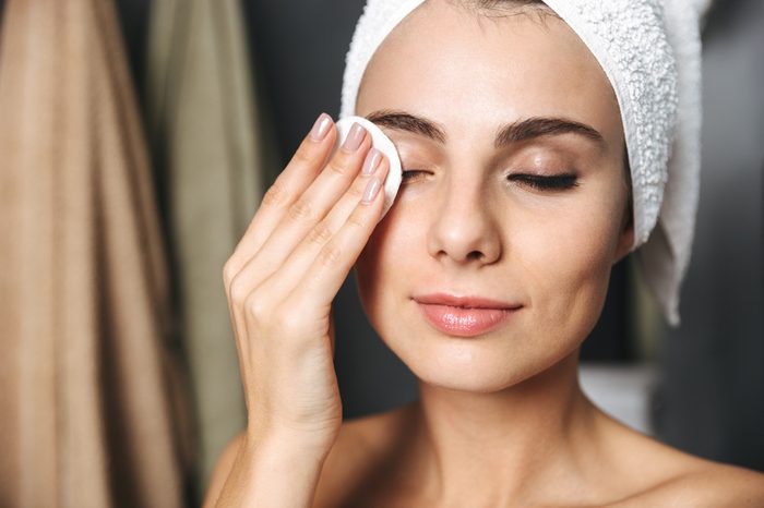 Beautiful young woman wrapped in bath towel standing at the bathroom, removing makeup with a cotton pad