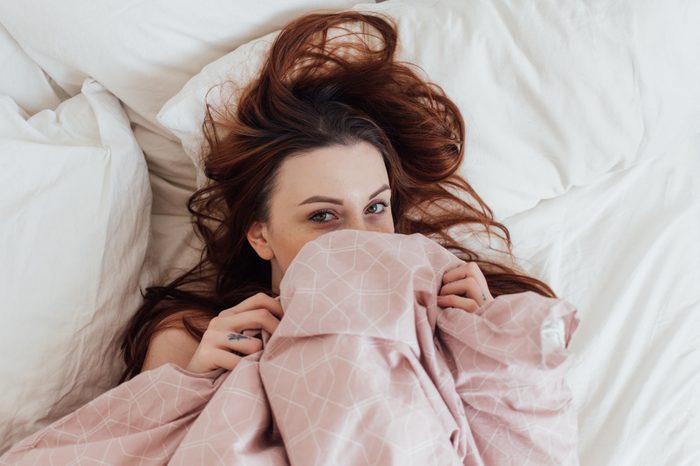 Cute and beautiful, pretty lovely young woman or teenager, peeks out of duvet and bedsheets covers, lays in bed on early weekend morning, lazy and sensual hipster with tattoos