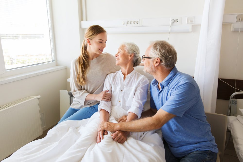 medicine, support, family health care and people concept - happy senior man and young woman visiting and cheering her grandmother lying in bed at hospital ward