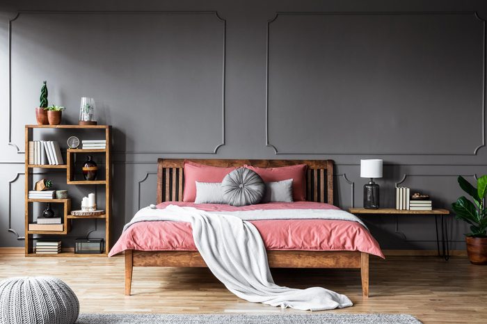 A large bed in a spacious, dark bedroom standing between a shelf with books and ornaments and a bedside table with lamp