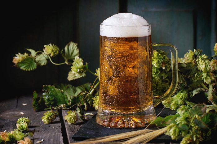 Mug of fresh draught lager beer with foam on black slate, served on old wooden table with green hop and ears of barley. Dark rustic style