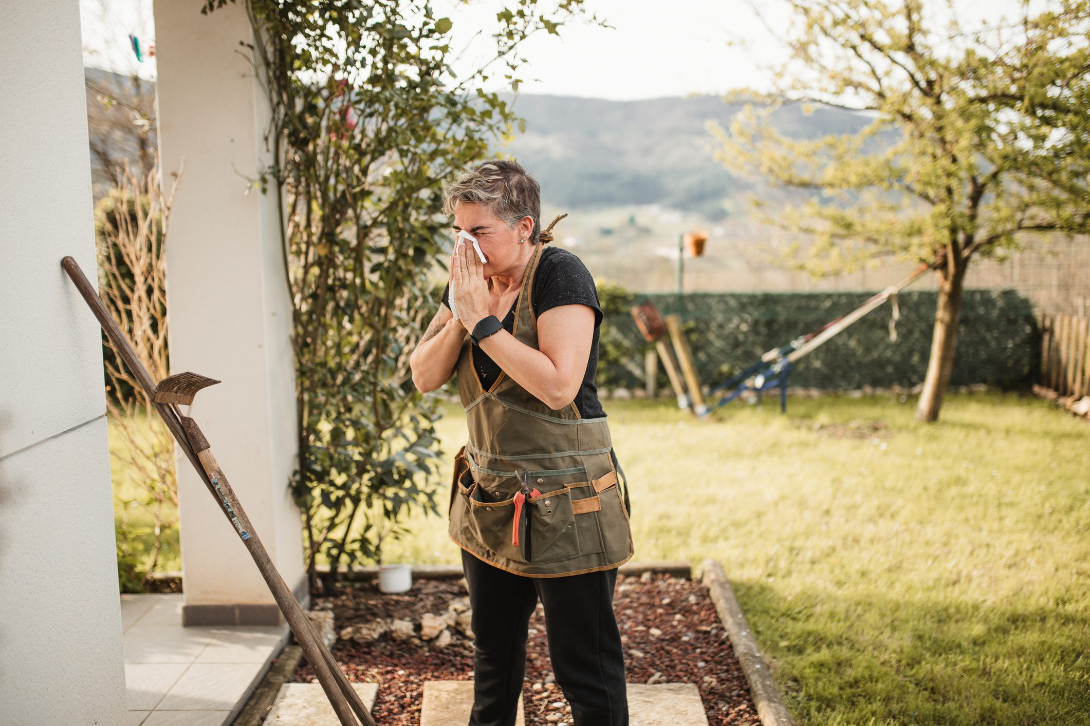 adult woman suffering from spring allergy and blowing a runny nose with a tissue while working in the garden