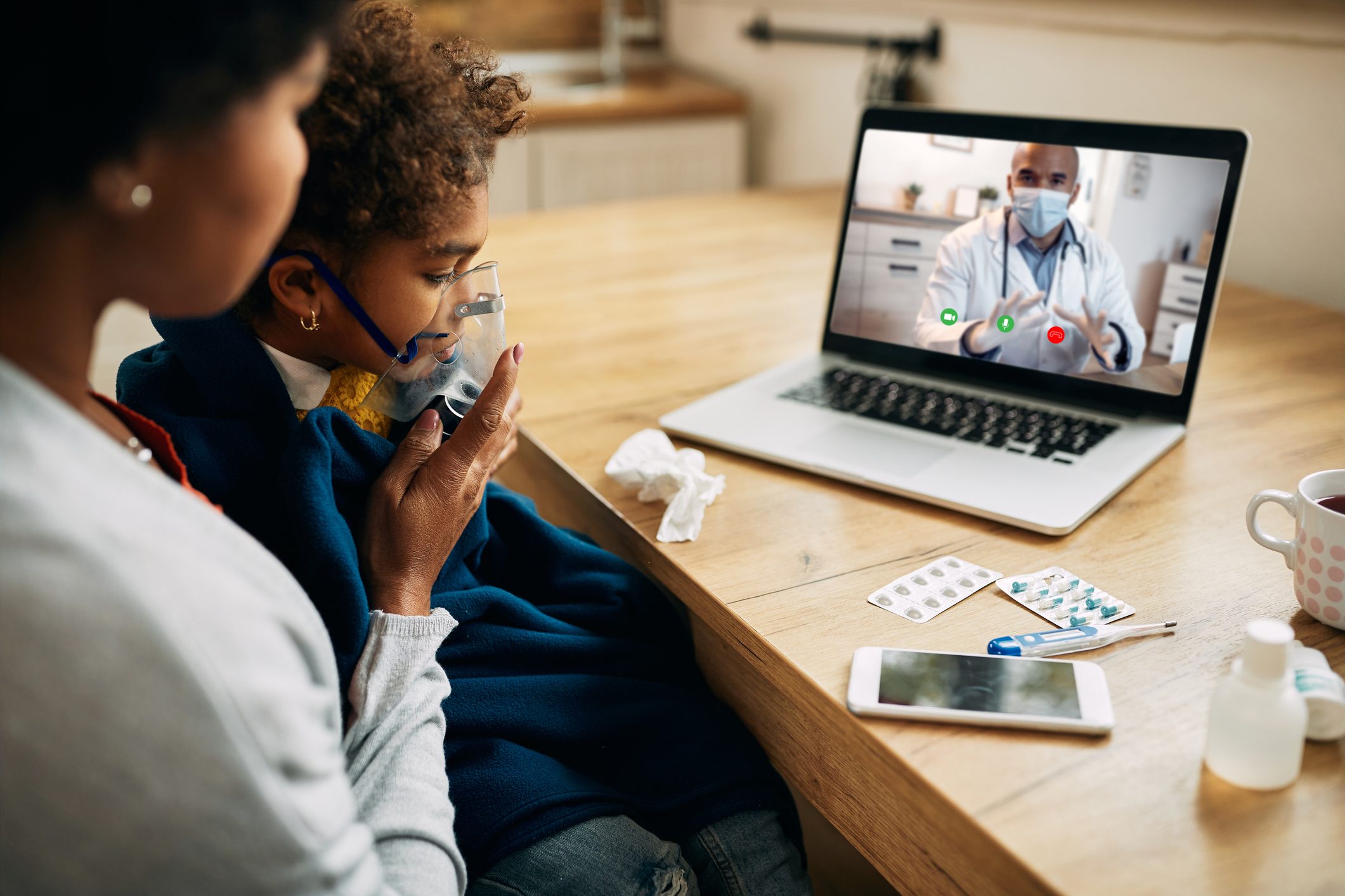 Black pediatrician talking via video call to the mother who is giving her daughter inhaling therapy at home.