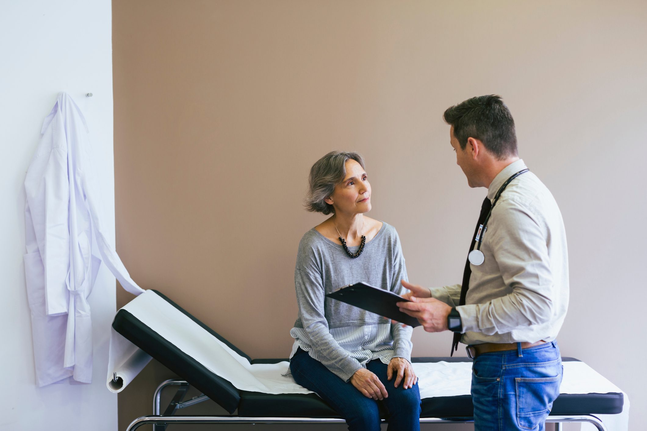Senior patient talking with doctor while sitting on bed against wall at hospital