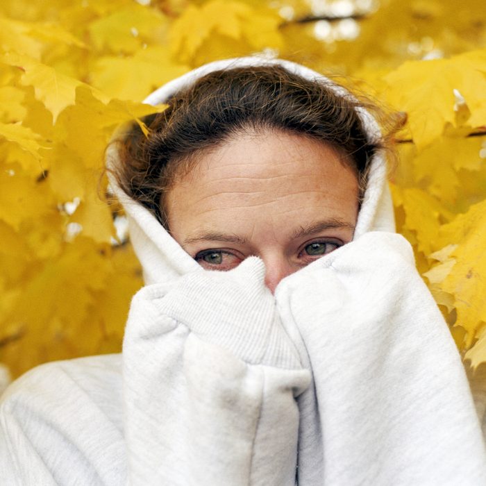 Woman wearing hooded sweatshirt pulled over her face with yellow autumn leaves behind her
