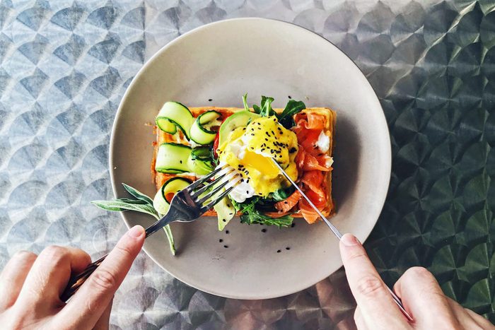 using a knife and fork to cut a waffle while Eating brunch with waffle, avocado, cucumber, salmon and poached egg, personal perspective