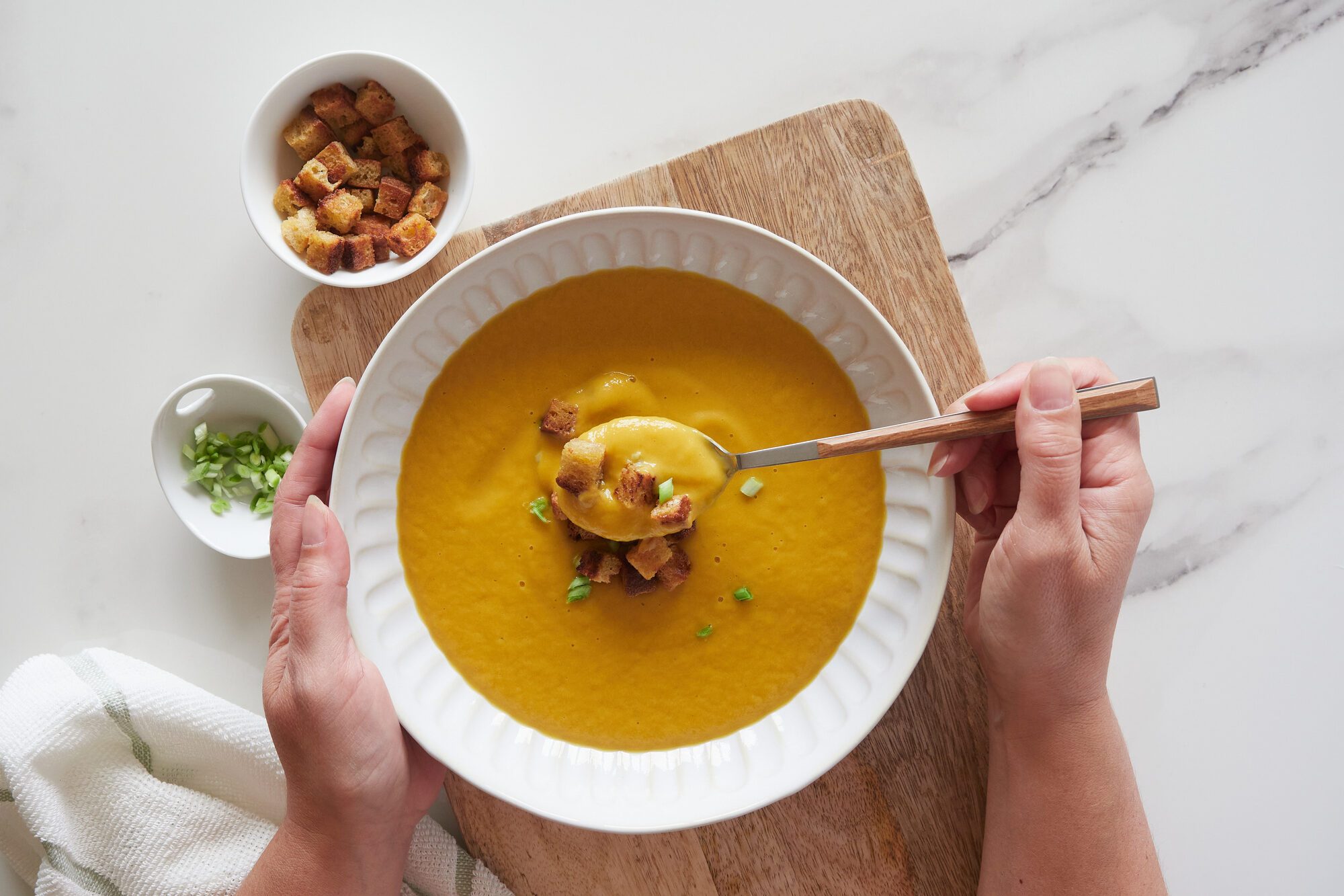 Top view of hand holding a spoon of pumpkin vegetable soup in a plate with bread croutons and spring onion topping on wooden board and marble kitchen countertop.