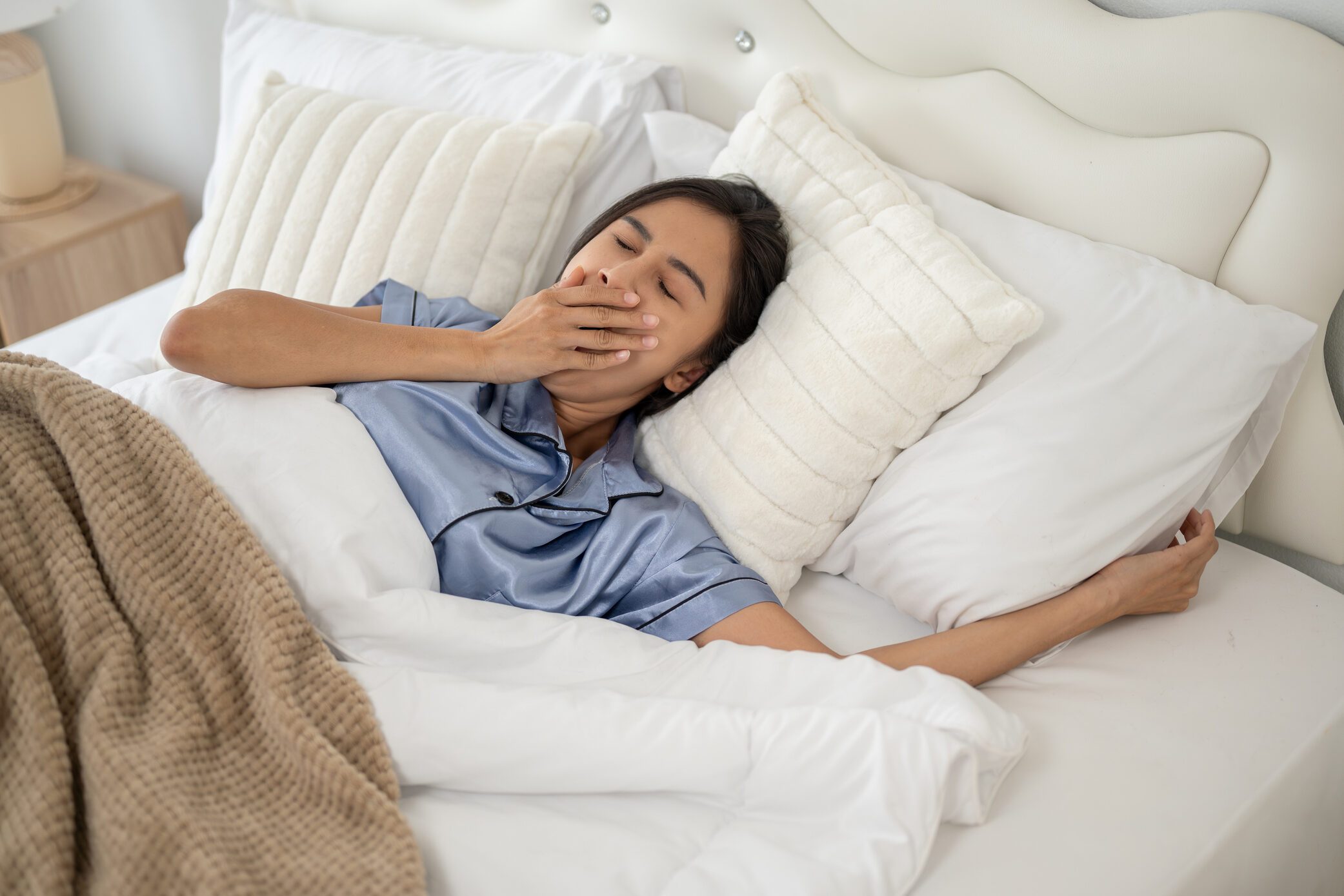 A sleepy and carefree Asian woman is yawning after waking up in the morning on her comfy bed.