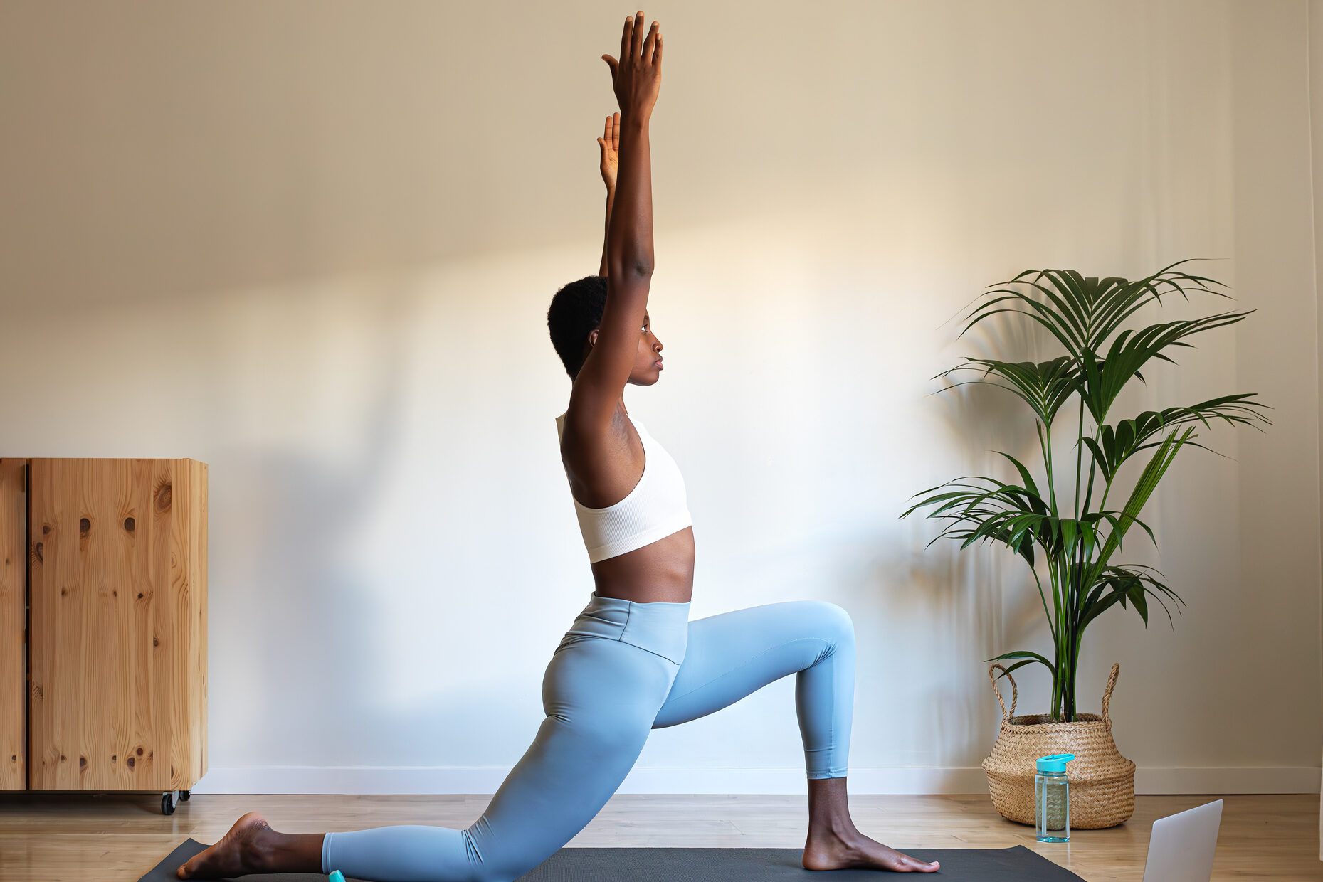 Young African American woman doing low lunge yoga pose at home with online tutorial using laptop. Anjaneyasana home yoga practice.