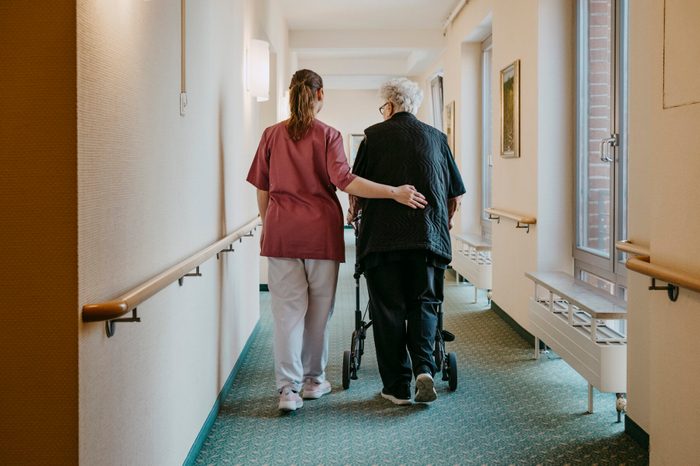Rear view of female caregiver assisting senior woman walking with mobility walker in corridor